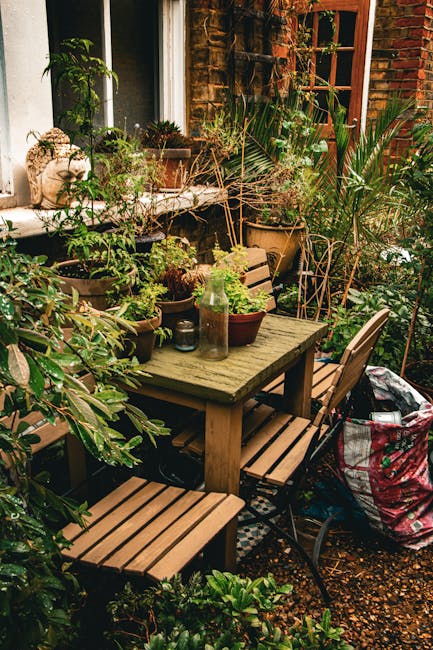 A cozy small garden terrace featuring a weathered wooden table surrounded by matching benches and chairs, set against a brick wall with a window. The table is decorated with potted plants, including s