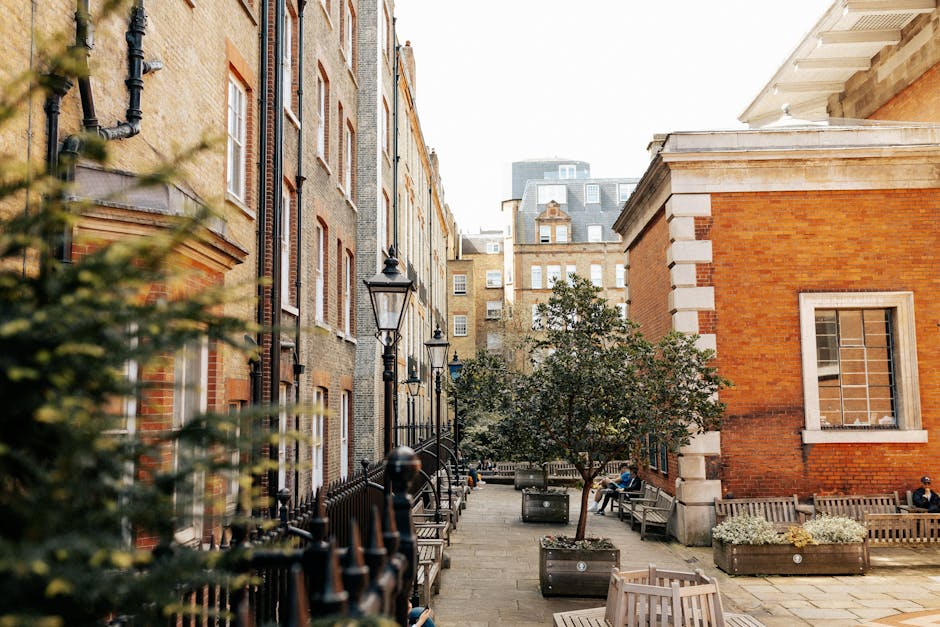 A modern urban courtyard featuring a neatly maintained garden bed with lush green shrubs and a small tree with textured leaves, contrasted against the sleek, curved glass facades of contemporary offic