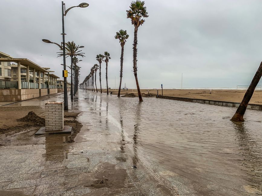A coastal promenade during overcast weather, slightly flooded from recent storm and flood damage. The paved walkway is wet with water pooling and small ripples across its surface, reflecting the gray 